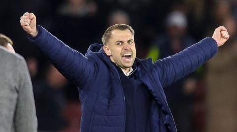 A close-up of Tonda Eckert holding his arms out in celebration wearing a blue puffer coat over the top of a blue zip-up jacket and a white collared top