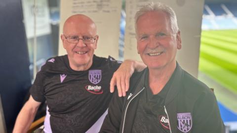 John Homer and Ally Robertson sit side by side smiling and both wear black t shirts with the west brom badge on