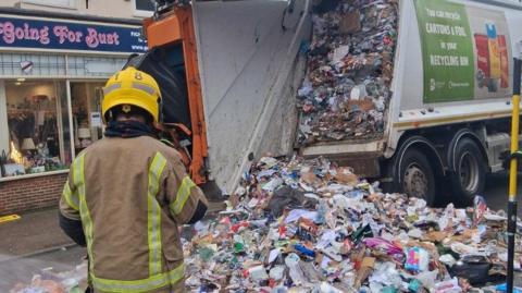 The contents of a waste collection truck spilled across a street. A worker wearing a helmet is standing at the rear of the truck.