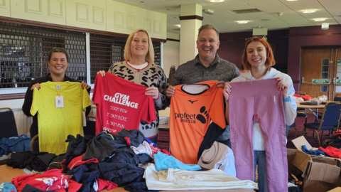 Three women and one man hold up sports shirts and leggings while standing in a community hall. There are clothes all over the tables.