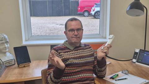 Andrew Fowler - a man sitting looking at the camera with a landline in his hand in an office. He is looking glum
