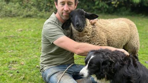 A man, sheep and a collie dog are huddled on a field. The man is hugging the sheep and is resting a hand on the dogs back.
