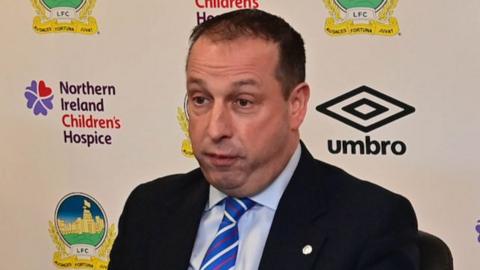 Roy McGivern speaks at a press conference. He has short black hair and is wearing a dark suit, light blue shirt and red, white and blue tie. He is sitting in front of a billboard with the Linfield FC crest, the Umbro logo and the NI Children's Hospice logo.