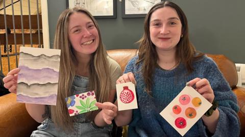 Poppy Barrett (left) and Hannah Broughton (right) are sitting on a brown leather sofa, each holding up small pieces of colourful artwork. The pieces include abstract purple waves, bright green leaves with a pink flower, a red pomegranate, and several circular fruit designs, possibly tomatoes of different colours. Behind them is a dark green wall with framed art.