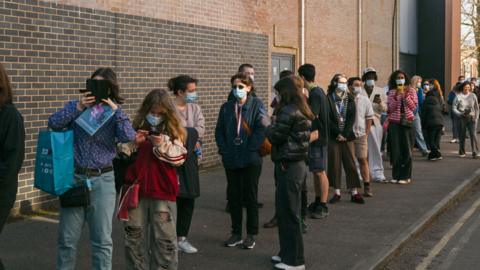 Students, some wearing masks, queuing outside a building
