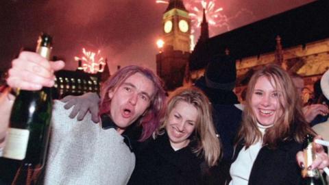 Three people with champagne bottles ring in the year 2000 infront of fireworks and Big Ben