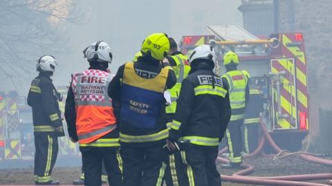 Firefighters, a safety officer and hazmat advisor stand in a group wearing high-vis firefighting attire and hard hats. The air around them is smoke-filled and multiple fire engines can be seen in the background.