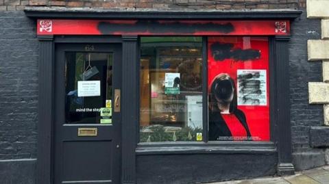 A black shop front has black spray paint on its windows and red shop sign. There is spray paint covering a window decal of Julia Buckley's face.