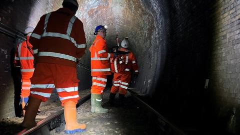 Workers in orange protective clothing, helmets and torches around their heads are surveying a dark railway tunnel at Whitehaven.