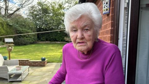 Brenda Catterall standing in her garden. She is an elderly woman and has white hair. She is wearing a bright, purple top. There is a patio with a chair and table on it and trees and a lawn are in the background. 