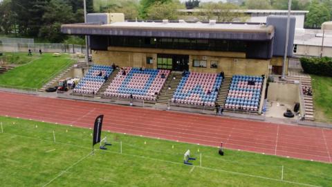 A picture taken from bird's eye view of an athletic stadium. You can see the stand which is red and blue in colour with the letters 'P', 'O', 'W' and 'S' spelt out on the seats. A running track can be seen in front of the stand.