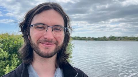 A man smiles as he stands on the shore of a large lake. He has long, dark-brown hair and a beard and wears a black jacket, blue shirt and rectangular glasses. The lake looks grey under a cloudy sky. The banks are covered in green foliage.