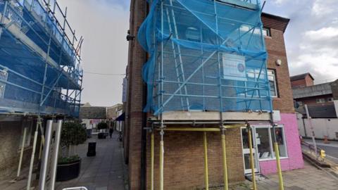 A brick building, which has part of its facade painted pink, with scaffolding around it.