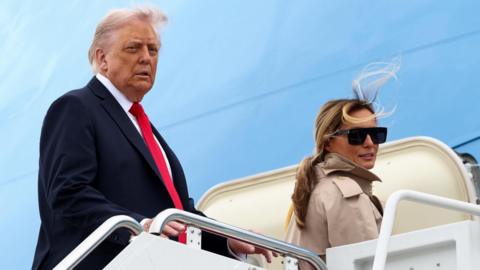 Donald Trump standing on steps leading to a plane, wearing a dark suit with a white shirt and red tie, alongside Melania Trump who is smiling slightly and wearing dark sunglasses and a tan coat with turned up collar