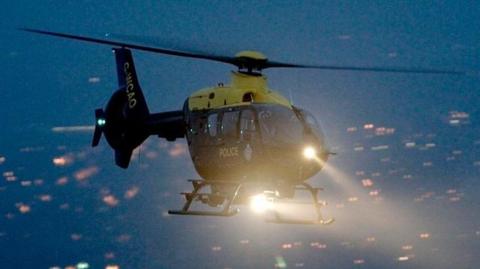 A police helicopter flying over a residential area at night. Lights below indicate many houses. There are two search lights on the helicopter which are brightly lit.