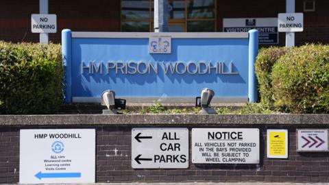 A blue sign marking the entrance to HM Prison Woodhill is mounted in front of a brick building, surrounded by hedges and multiple notices, including “No Parking” and directions to visitor and parking areas.