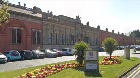 A red and yellow brick building with cars parked in the front. A grass area is planted with flowers and two small bushes. A sign reads Unilever Port Sunlight, Lever House.
