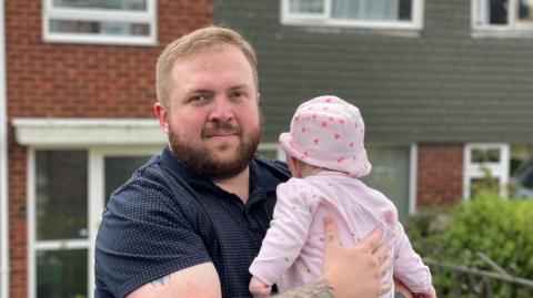 Bearded man wearing a blue polo shirt holding a baby girl wearing a pink baby grow and hat outside the their house in Telford