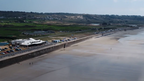 An aerial view of Watersplash Jersey. Watersplash is a white low level building on Jersey seafront. In front of it is a long, calm stretch of beach with the tide out. Watersplash is situated behind a sea wall. Cars can be seen parked in the nearby vicinity and there are green fields in the background. 