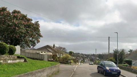 A street view of Conway Road in Falmouth. There is a garden with the grass to the left and houses on the right. There are cars parked on the road on the right side and pathways both sides.