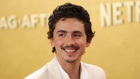 Timothée Chalamet smiling. He is wearing a white shirt and suit jacket. He has dark brown hair. The background behind him is gold.
