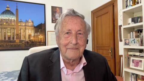 David Willey sat at home with a large picture of the Vatican behind him, wearing a suit and looking into the lens