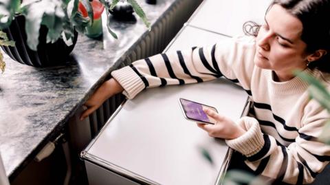 A high angle view of young woman wearing a white and black striped jumper who is checking the temperature of a radiator next to a window. She is looking at a heating app on her smart phone.