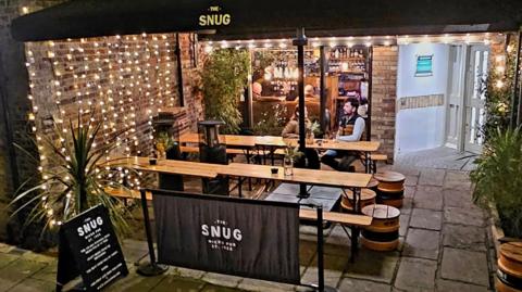 A couple sitting at a bench/table under a canopy. There are fairy lights covering a brick wall, various logos for The Snug, including on an A-board, which appears to be on a pavement. Inside the pub there is a bar and people sitting in chairs next to the window.