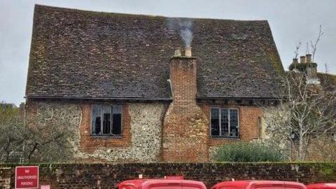 A medieval house is seen with smoke coming out of its chimney. It is made of brick and flint with a slightly sloping roof. Two window panes can be seen smashed.