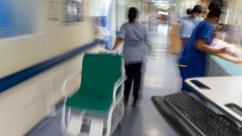 A blurred image of staff working in a hospital corridor wearing blue scrubs. One staff member is pulling a green wheelchair.