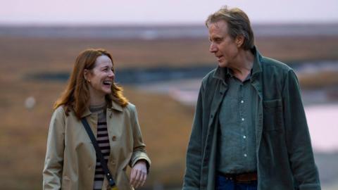 A screengrab of the film showing Rhys Ifans and Laura Linney looking at each other. Linney laughs while Ifans smiles at her. She wears a beige trench coat on top of a striped top, with her long brown hair reaching her shoulders. He wears a dark blue overshirt, with a blue shirt underneath and has short dark hair. 
