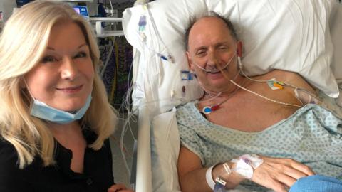 Mike is sat in a hospital bed with lots of tubes coming out of his right hand and chest and around his face. He is smiling at the camera. His blonde wife, Anna, is sat next to him also smiling at the camera. 