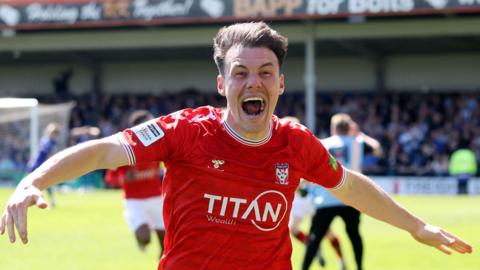 Ryan Fallowfield with his arms out wide in celebration and a huge smile on his face as York score the promotion winning goal at Rochdale 