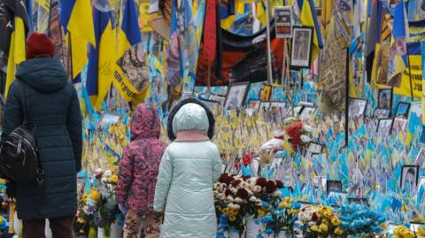 A woman and two young girls walk near a makeshift memorial to the fallen Ukrainian servicemen and international volunteers, in Independence Square in Kyiv, Ukraine. The monument is covered in flags and photographs. Flowers adorn the foot of the structure.