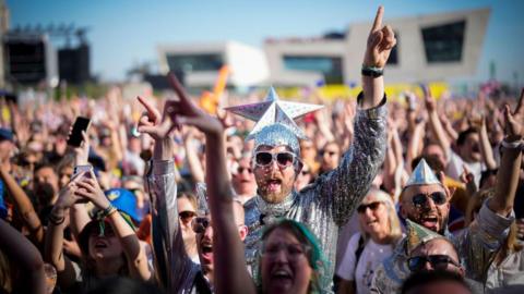 Eurovision fans watch the 2023 Eurovision Song Contest final on a giant screen in host city Liverpool. Many fans are dressed up, including the man in the centre of the photo who wears a silver jumpsuit and sunglasses and wears a silver cap with a giant silver star on the top.