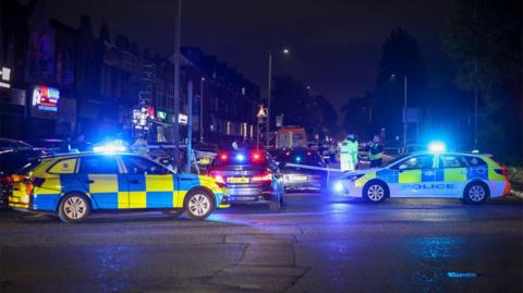 Police vehicles parked by police tape at Dickenson Road in Longsight.