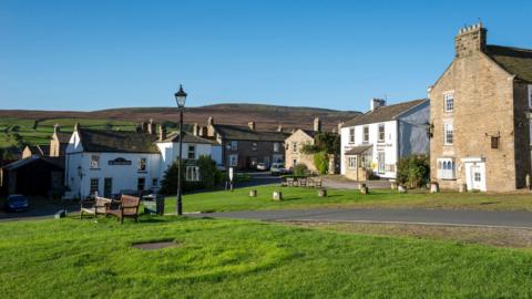A view of a village in the Yorkshire Dales, with traditional whitewash and red brick buildings, set around a village green. A heather-covered hill can be seen in the background. 