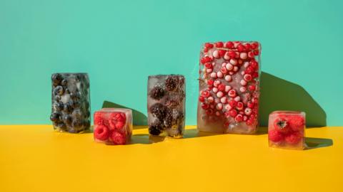 Five blocks of frozen fruit in ice, l-r: medium sized block of blueberries, small sized block of raspberries, medium sized block of blackberries, large sized block of red currents and small sized block of strawberries. They sit on a bright yellow surface with a turquoise background. 