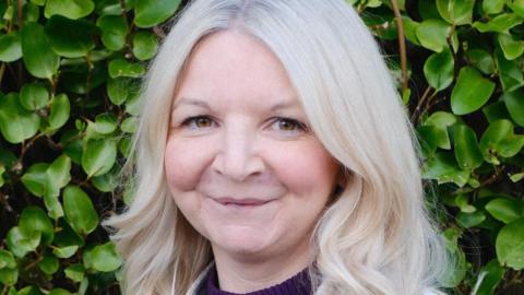 Naomi McBurney looking straight into camera, she is standing in front of a green leafed bush