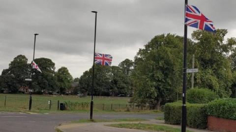 Union jack flags placed on lampposts in a street that has greenery and bushes and trees in the background