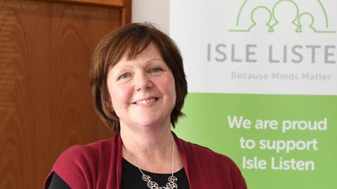 Andrea Chambers, a woman with short brown hair, smiles and wears a red cardigan as she stands in front of a poster that says Isle Listen.