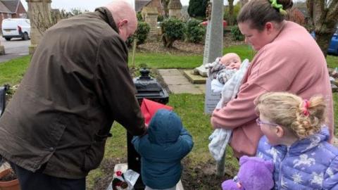 A family gather to post a card in a red envelope at the black post box in a graveyard. A little child in a blue hooded coat is helped by a man on the left to put the card in, while a woman on the right holding a baby and a little girl in a purple coat standing on the right watching.