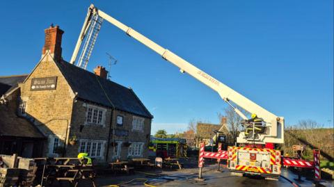 A traditional country pub can be seen with picnic-style tables laid out around the front of the building. To the right, a large crane is being operated by a firefighter. The arm of the crane is extending up and over the pub. From the end of the crane's arm, a ladder descends down to the pub's roof. A fire engine can be seen tucked behind the pub.