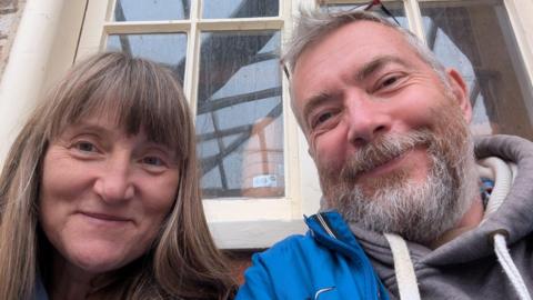A woman is lookng at the camera smiling, standing shoulder to shoulder with a man who is also smiling. Both are looking at the camera, and behind them is a window with stacked plant pots appearing through the glass.
