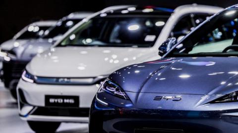 A row of four BYD cars are displayed at the 46th Bangkok International Motor Show in Bangkok, Thailand. The sedan in the foreground is a blue vehicle with sharp, aggressive looking headlamps. The car behind it is a larger vehicle, the BYD M6.