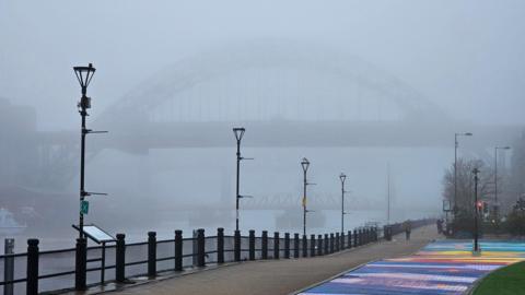 The Tyne Bridge pictured from the Newcastle side in the fog. In the front of the photo is a multi-coloured rainbow path on the quayside. Below the Tyne Bridge you can see the outline of the Swing Bridge and the High Level Bridge. There are multiple lamp posts on the side of the river.