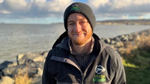 A man standing in front of Belfast Lough. He is wearing a black and grey rain jacket with an Ulster Wildlife logo on it. He is also wearing a black hat. 