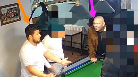 A screenshot from CCTV footage showing a group of men gathered around a pool table in a pub. The man on the left, with dark hair and wearing a white t-shirt, has been highlighted with an orange arrow pointing at his head. The second man on the right, who is bald and wearing a black t-shirt and brown jacket, is highlighted with a pink arrow. The other people in the image have been intentionally blurred out.