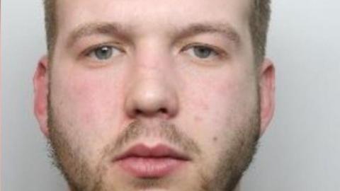 A custody picture of a man with light brown hair and a beard staring straight ahead. He is wearing a grey top. 