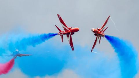 The Red Arrows perform at RAF Fairford during the Royal Military Air Tattoo on July 15, 2023 in Fairford. Two red aircraft trailing blue smoke perform a close pass with a third aircraft with red smoke in the background.